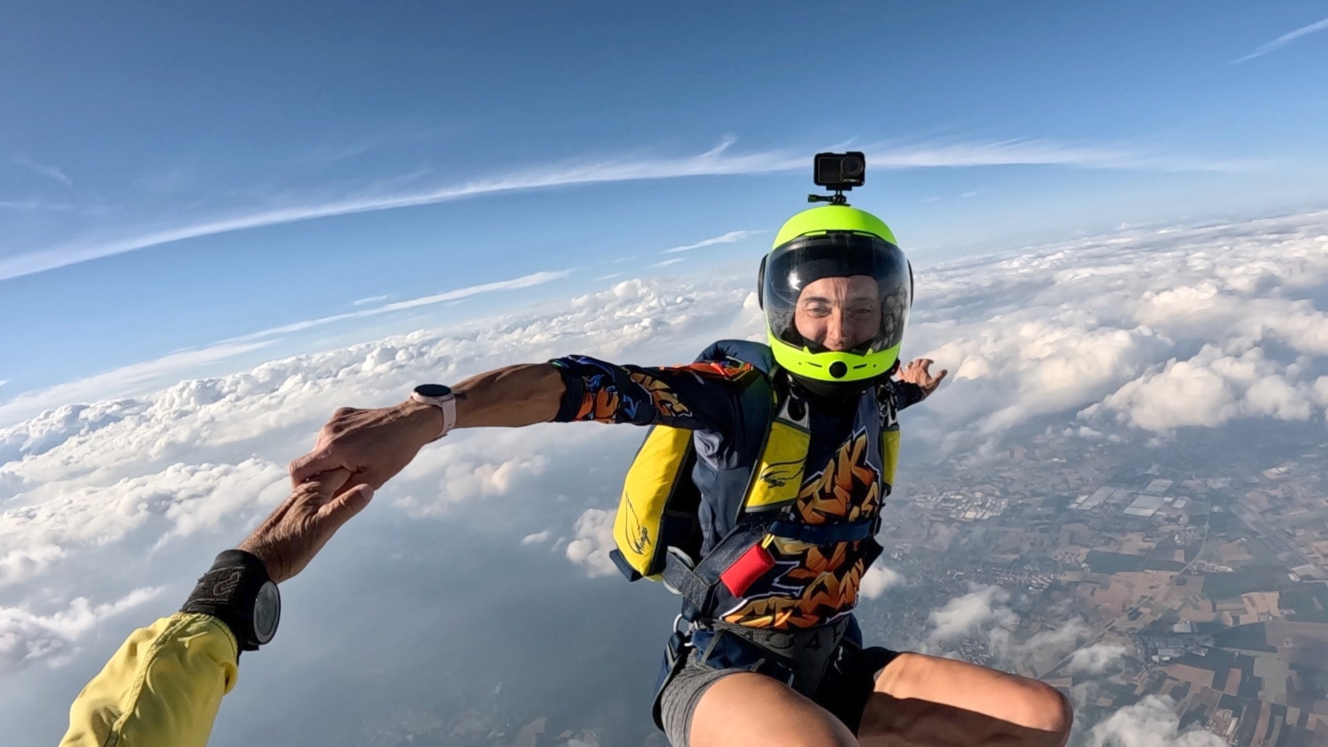 A skydiver in a black, orange, and yellow called Graffiti Fck-Theground jersey with a bright yellow helmet and backpack is freefalling above the clouds, reaching out to be held by another person wearing a yellow jacket.