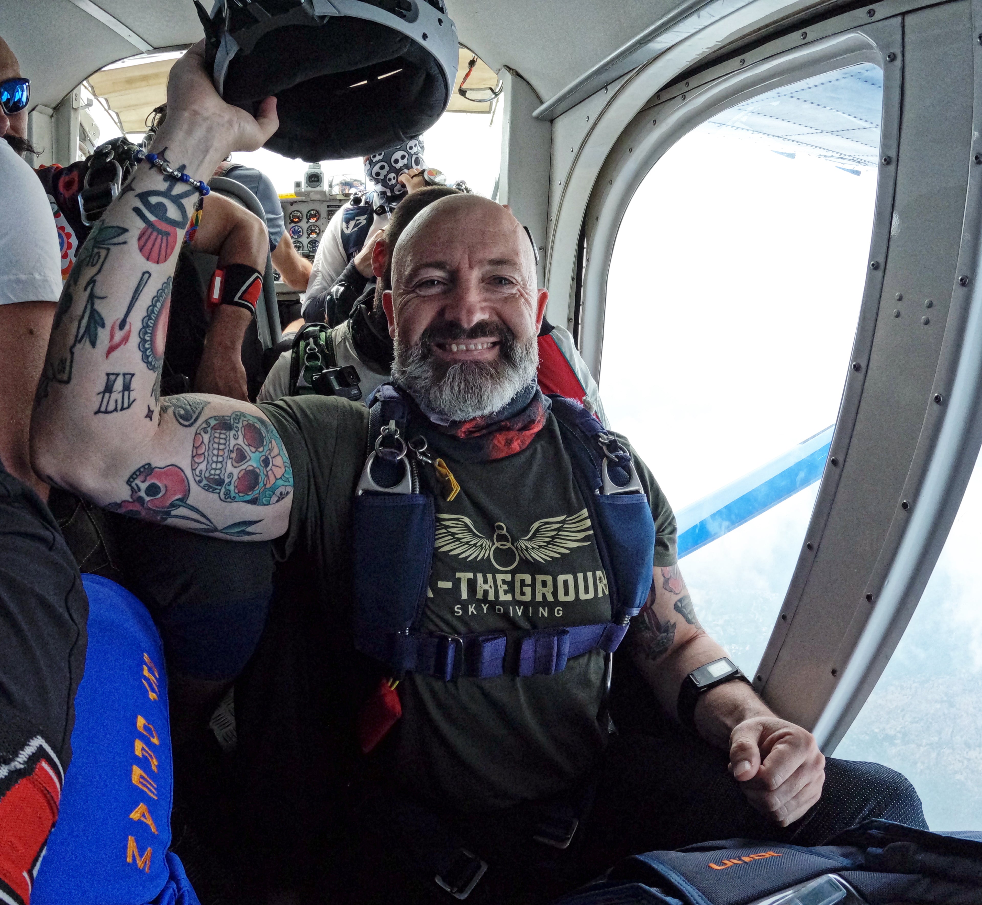 A smiling skydiver wearing a dark olive green t-shirt FCK-THEGROUND brand, a blue harness, and holding a helmet inside the cabin of a plane.